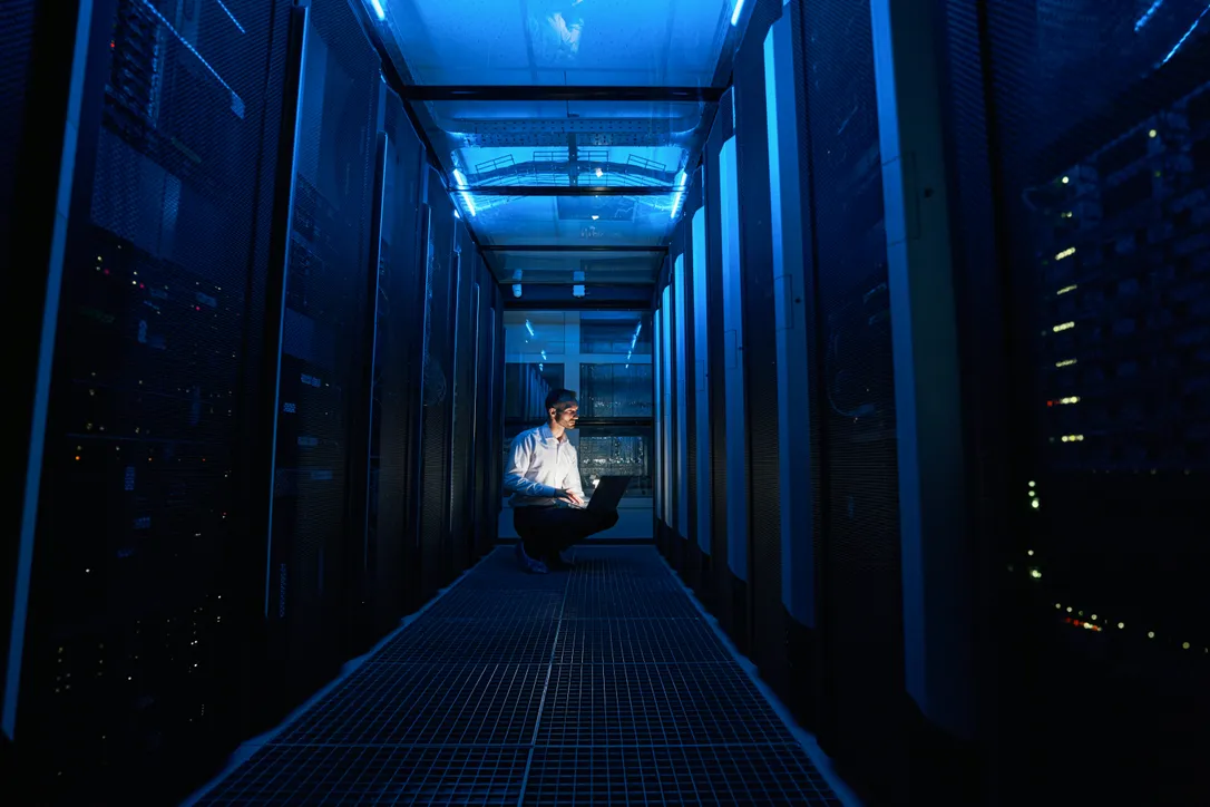 Technicien informatique travaillant sur un ordinateur portable dans un couloir de centre de données avec serveurs éclairés en